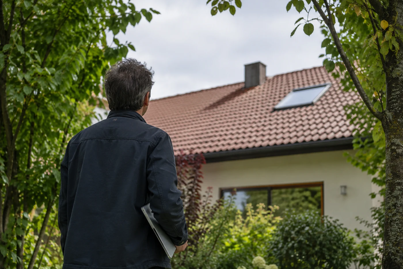 Person betrachtet ein Hausdach zwischen Gartenbäumen